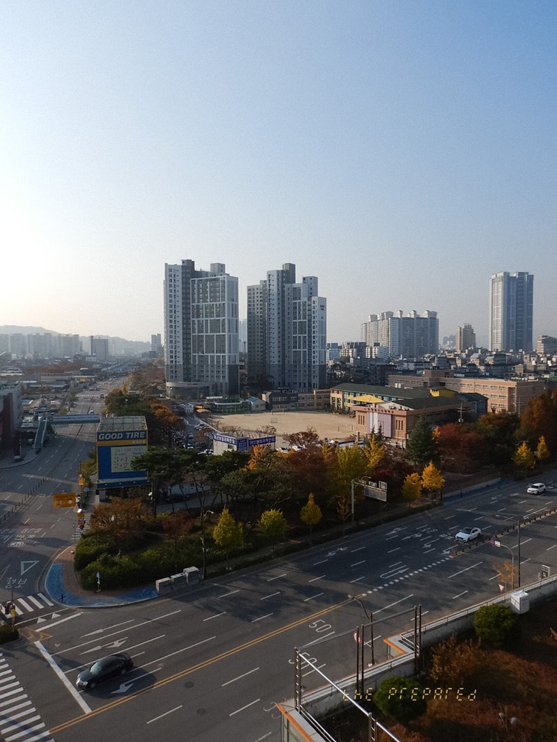 이 이미지는 한국 충청남도 아산의 도시 풍경을 보여줍니다. 이 지역은 아파트 건물, 거리 및 기타 건물이 있는 주거 지역으로 보입니다. 이미지 정보는 다음과 같습니다. 위치: 아산시, 충청남도, 대한민국. 전반적인 관점: 도시 풍경 주요 피사체: 고층 아파트 건물, 도시 거리, 자동차 이미지에 포함된 내용: 도시 건물, 나무가 늘어선 거리, 도로, 자동차,