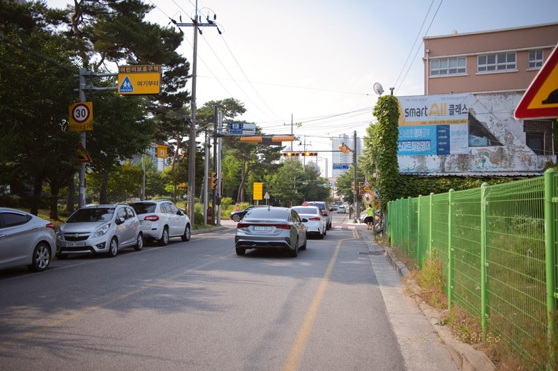 이 이미지는 낮 시간대에 촬영된 아산시 충청남도의 거리를 보여줍니다. 도로를 따라 자동차가 줄지어 있고, 오른쪽에는 초록색 울타리가 있습니다. 표지판에는 