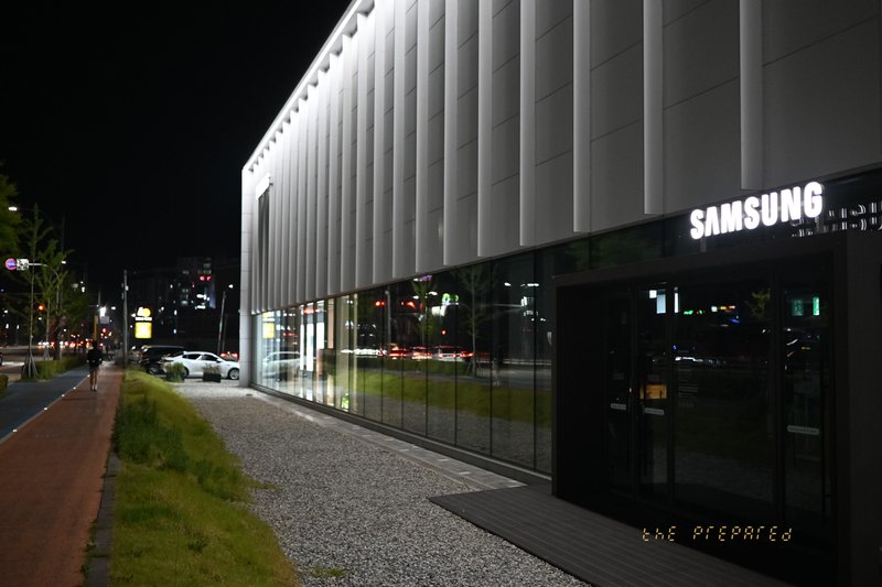 저녁에 건물 외부에서 찍은 삼성 매장 사진. 건물의 외벽은 회색과 흰색 줄무늬로 덮여 있으며 큰 삼성 로고가 건물 앞면에 있습니다. 매장 앞에는 포장된 보도가 있습니다.