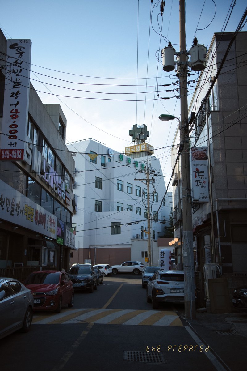 아산시 도심의 좁은 골목길을 담은 사진. 전봇대와 복잡하게 얽힌 전선이 보이고, 양옆에는 상가 건물이 늘어서 있습니다. 골목길 안쪽에는 하얀색 건물이 보이며, 골목길에는 차량 몇 대가 주차되어 있습니다.