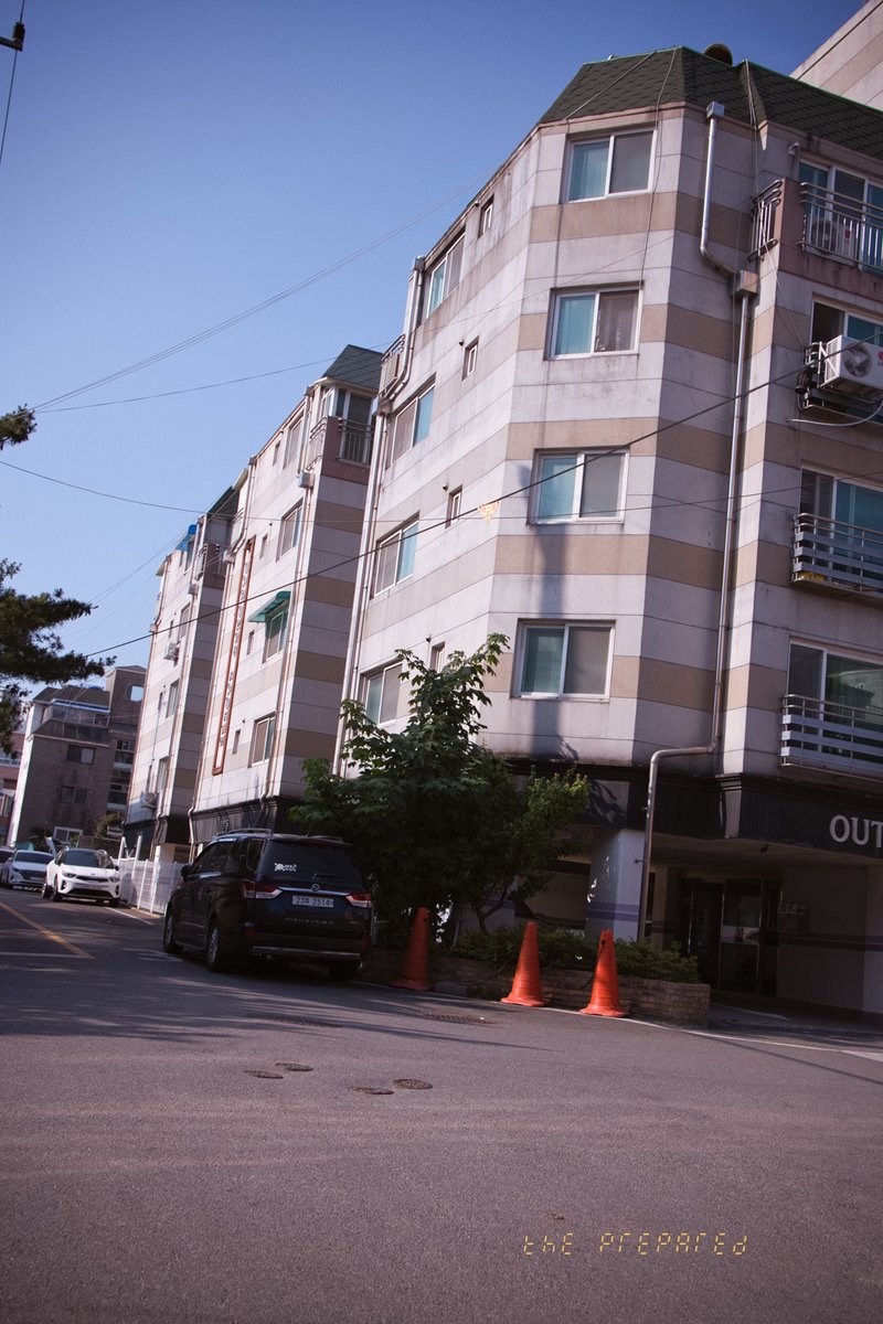 흰색과 회색의 아파트 건물입니다. 이미지에는 도로, 자동차, 3개의 주황색 교통 콘과 텍스트의 일부가 있습니다.