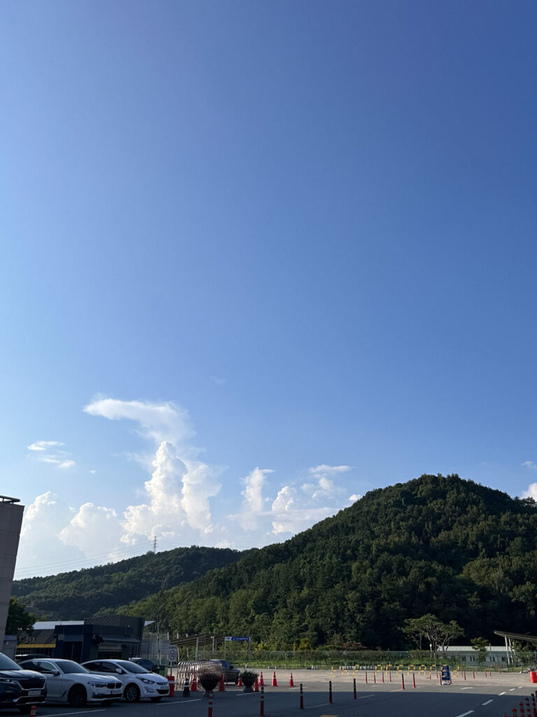 자동차 몇 대가 보이는 산과 하늘 사진. 이미지는 울창한 녹색 나무로 덮인 언덕의 넓은 풍경을 보여줍니다. 언덕 앞에 넓은 주차장이 있으며, 여러 대의 자동차와 주차 장애물이 보입니다. 사진은 맑은 푸른 하늘과 흰 뭉게구름이 떠있는 것을 강조합니다.