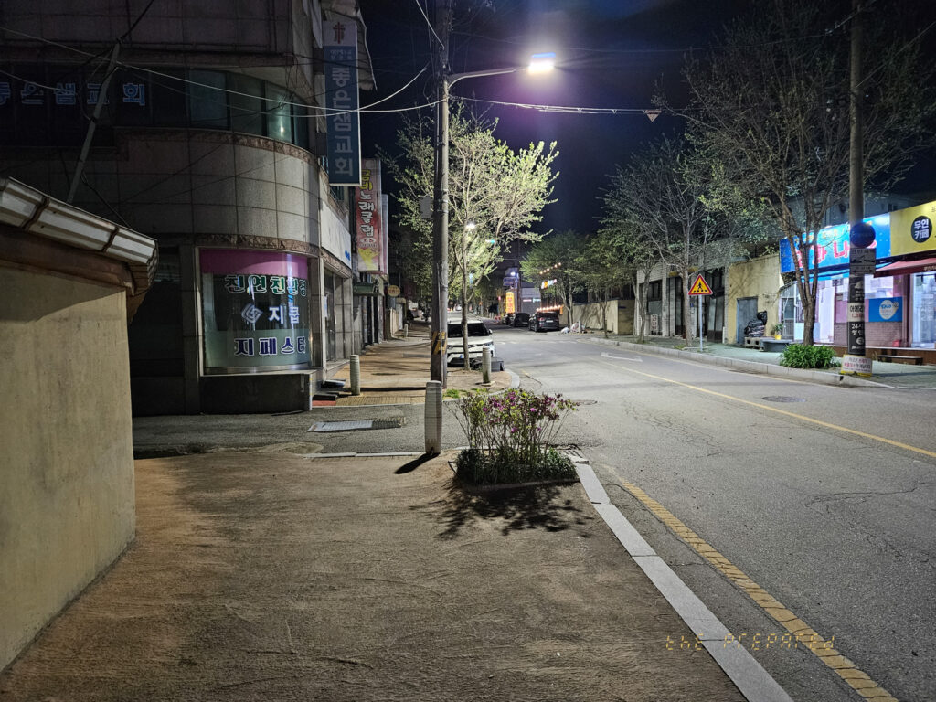 이 이미지는 저녁에 한적한 거리를 보여줍니다. 양쪽에는 건물이 늘어서 있으며, 오른쪽에는 가로등이 켜져 있고 나무 몇 그루도 보입니다. 아스팔트 도로는 거리를 따라 구불구불 이어져 있으며, 거리를 따라 다양한 표지판과 조명이 흩어져 있습니다. 전반적인 분위기는 고요하며 인적이 드물고 차분합니다.