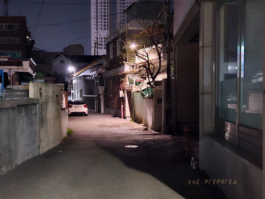 어두운 배경에 가로등 불빛이 골목길을 비추고 있으며, 건물과 주차된 차량이 보입니다.