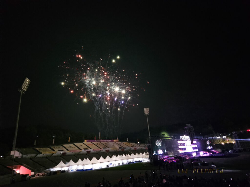 이 이미지는 어두운 밤에 불꽃놀이를 묘사합니다. 불꽃은 하늘에서 폭발하며 화려한 색상을 연출합니다. 아래에는 관중을 위한 좌석이 있는 축구 경기장이 있습니다. 경기장의 측면에는 경기장을 밝히는 두 개의 전등 기둥이 있습니다. 전반적인 장면은 축제적이며 흥분됩니다.