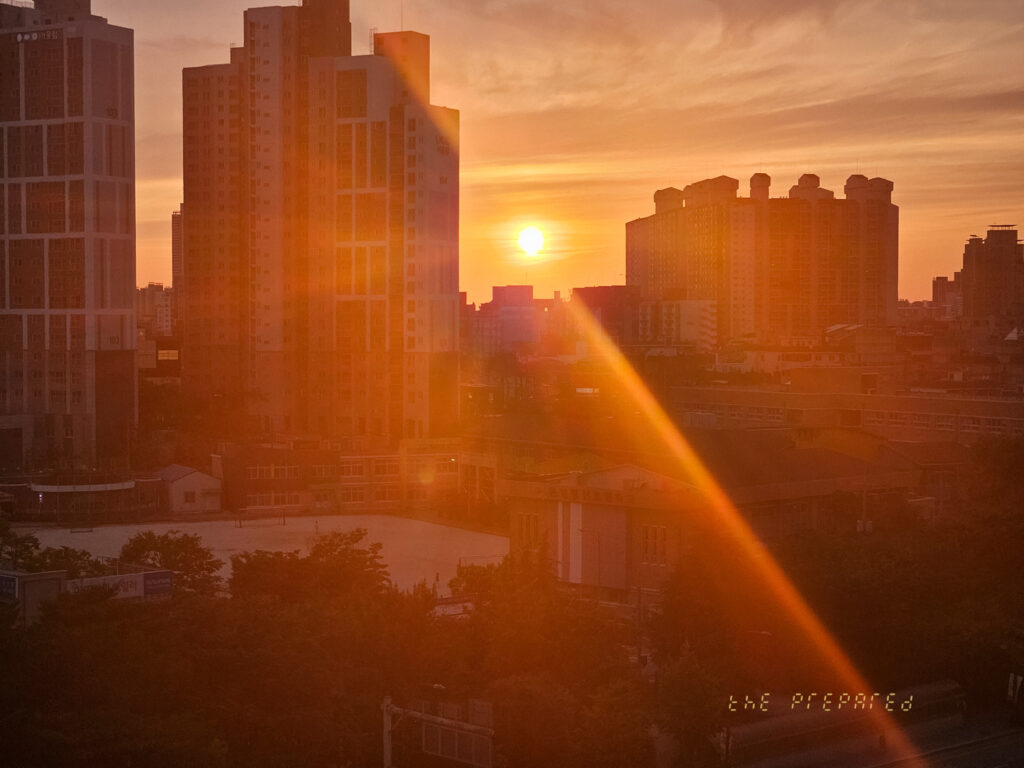 저녁 도시 풍경 사진입니다. 석양으로 물든 하늘과 도시 건물들의 실루엣, 그리고 태양이 만들어내는 강렬한 빛줄기가 인상적입니다