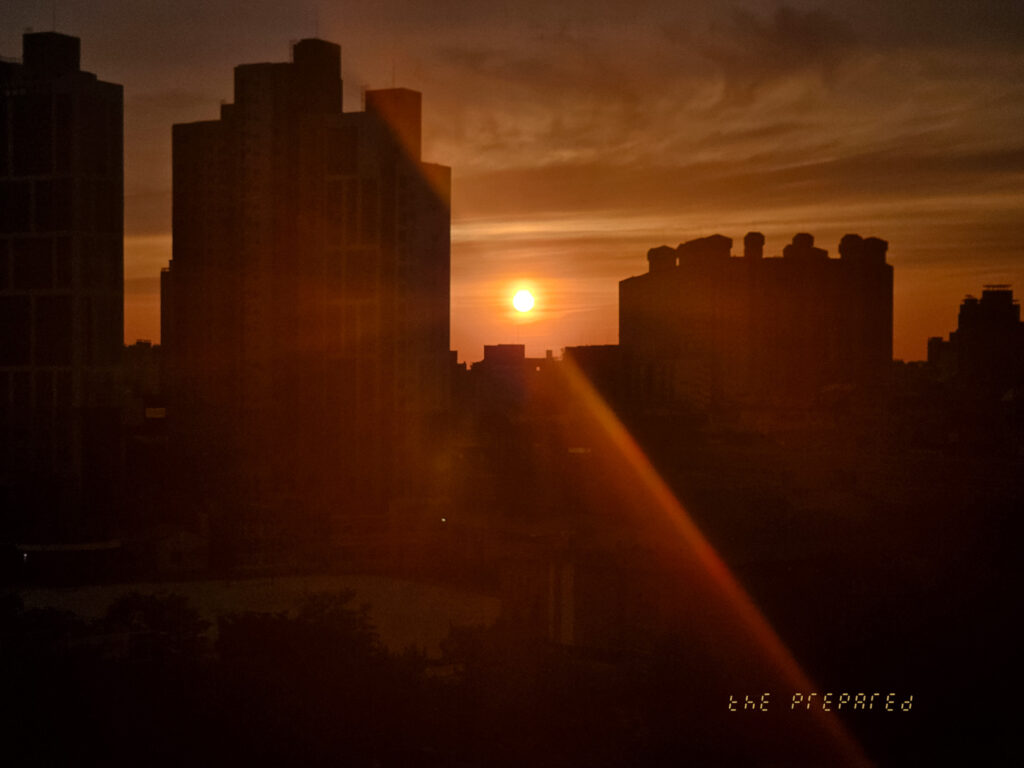 도심의 일몰 사진, 하늘은 석양으로 물들어 있고 건물은 어두운 실루엣으로 표현되어 있습니다. 태양빛이 건물을 비추며 강렬한 빛줄기를 만들어내고 있습니다.