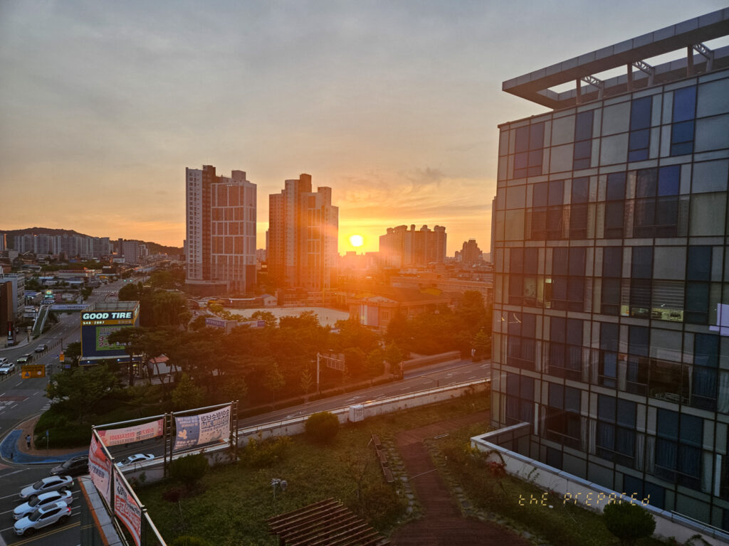 아산시에서 촬영된 일몰 사진입니다. 건물과 도시 풍경, 그리고 석양으로 물든 하늘이 보입니다. 오른쪽 건물 유리창에 햇빛이 반사되어 따뜻한 느낌을 줍니다.