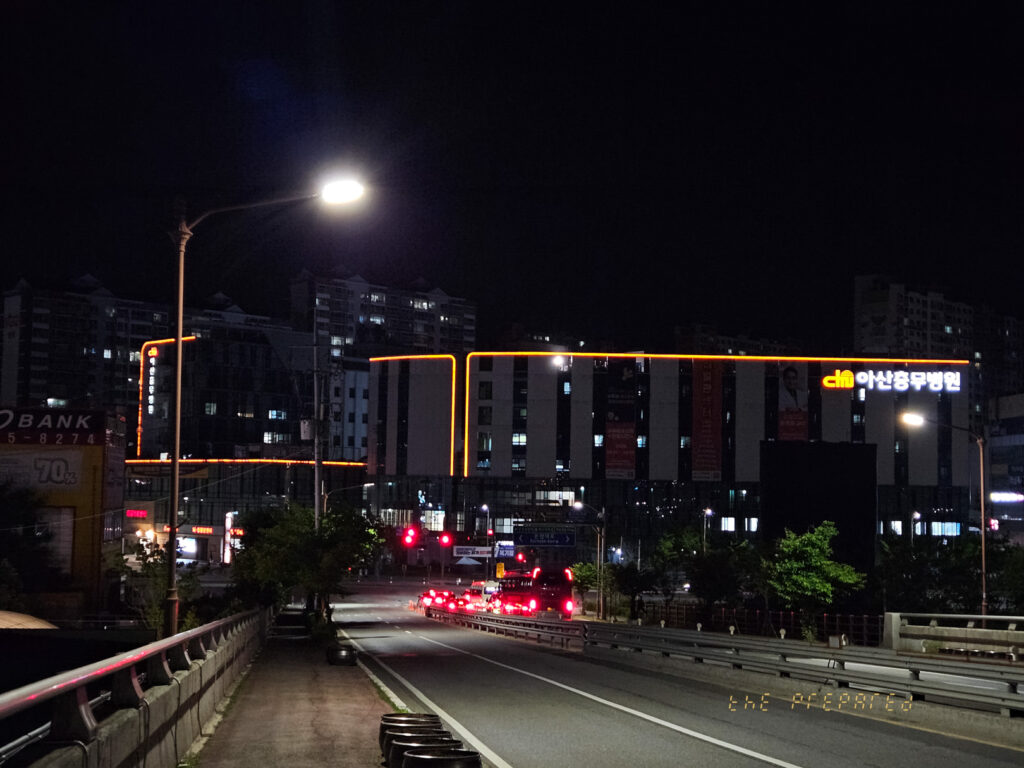 아산충무병원과 주변 도시 풍경이 어우러진 밤거리 사진. 병원 건물은 주황색 조명으로 강조되어 있으며, 거리의 차량과 가로등 불빛이 어둠 속에서 빛나고 있습니다.
