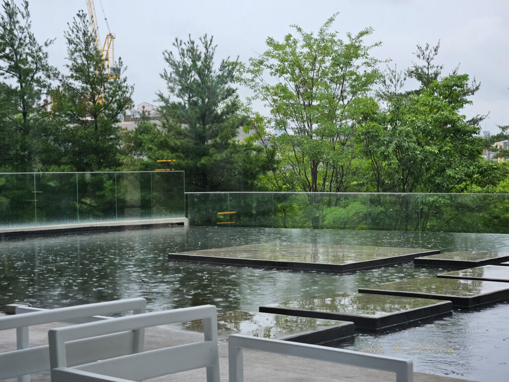 이것은 나무, 물, 그리고 몇 개의 정사각형 석판이 있는 멋진 야외 공간의 이미지입니다. 사진에는 물이 반사되는 넓은 표면과 나란히 놓인 여러 개의 사각형 돌판이 있습니다. 나무와 기타 식물은 무성한 배경을 만들어 냅니다. 현대적인 스타일의 좌석이 놓여 있어 세련되고 여유로운 환경을 조성합니다. 이미지에는 약간의 흐림 효과가 있습니다.