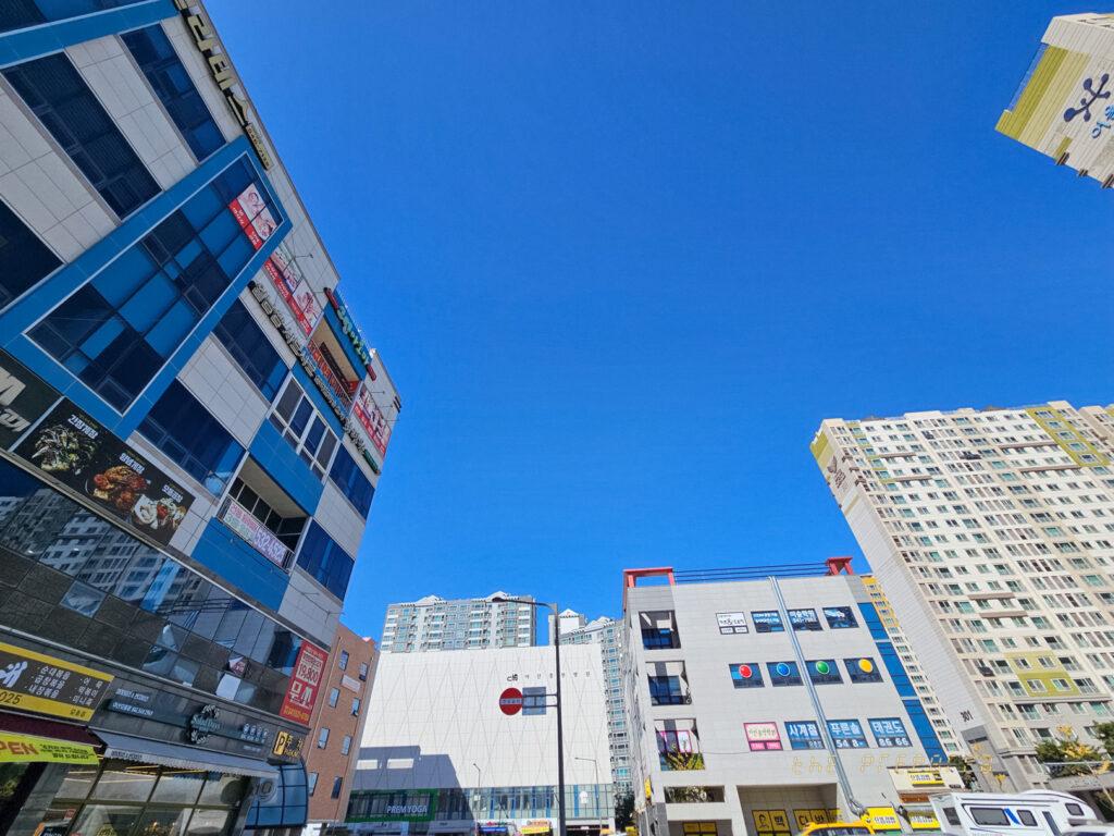 도시의 건물들이 보이는 맑은 하늘 사진. 왼쪽에는 파란색과 흰색 줄무늬가 있는 건물이, 오른쪽에는 다양한 색깔의 건물이 보입니다. 사진의 대부분은 맑고 푸른 하늘이 차지하고 있습니다.