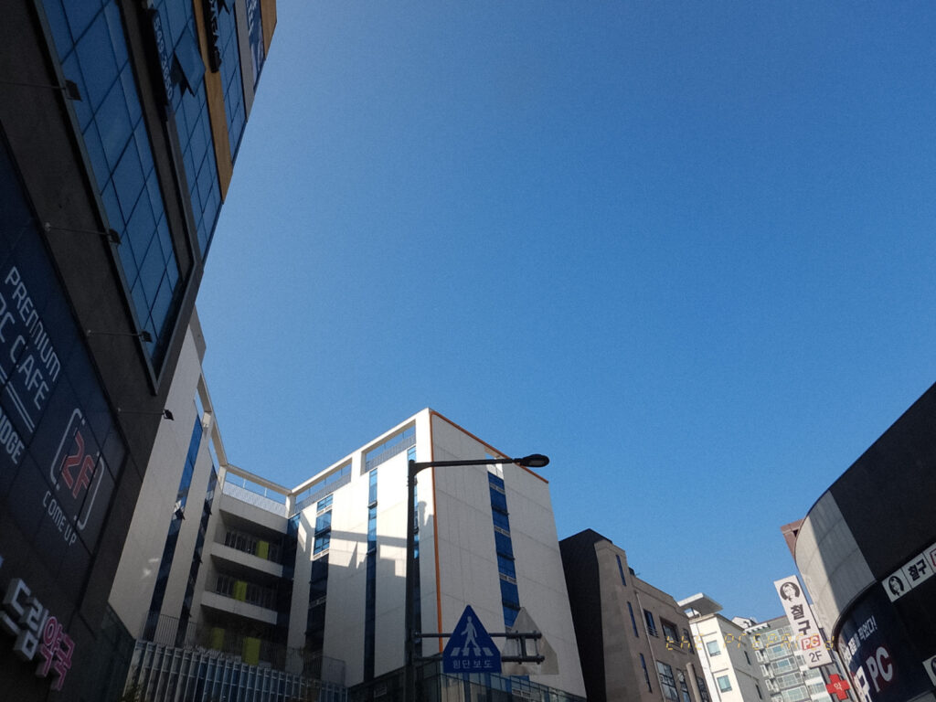 도심 건물들이 보이는 맑은 하늘 사진. 왼쪽에는 'PREMNIUM PC CAFE'라는 문구가, 오른쪽에는 '철구 PC'라는 문구가 적힌 건물이 있습니다. 위쪽에는 푸른 하늘이 넓게 펼쳐져 있습니다.