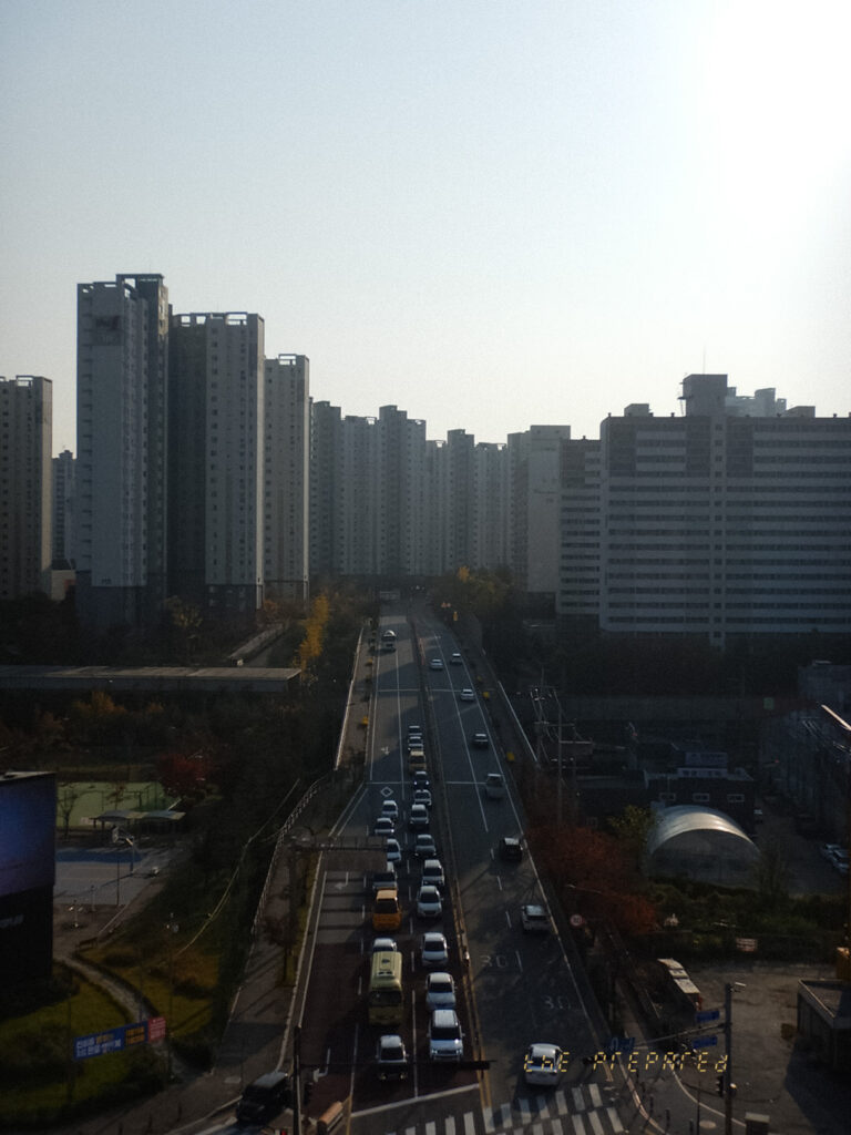 회색 고층 아파트 단지가 빽빽하게 늘어선 풍경 사진입니다. 길게 뻗은 아스팔트 도로 위에는 자동차들이 줄지어 달리고 있습니다. 도로 양옆으로는 푸른 나무들이 줄지어 서 있습니다.