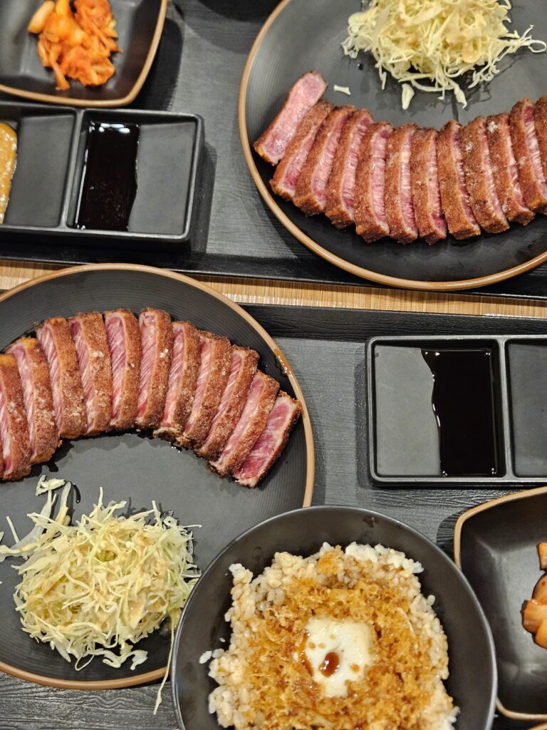 레스토랑 테이블 위에 놓인 규카츠 세트 사진입니다. 접시에 담긴 잘게 썬 규카츠가 보이며, 밥, 샐러드, 김치 및 소스도 함께 제공됩니다.