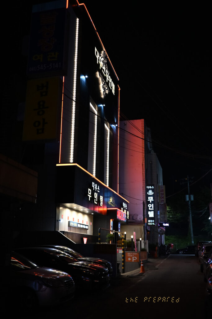 아산시의 어두운 밤거리를 담은 사진입니다. '다우스 무인텔'이라고 적힌 검은색 건물과 빨간색 여관 간판이 눈에 띄며, 길가에 주차된 자동차와 맞은편 건물의 조명이 보입니다.