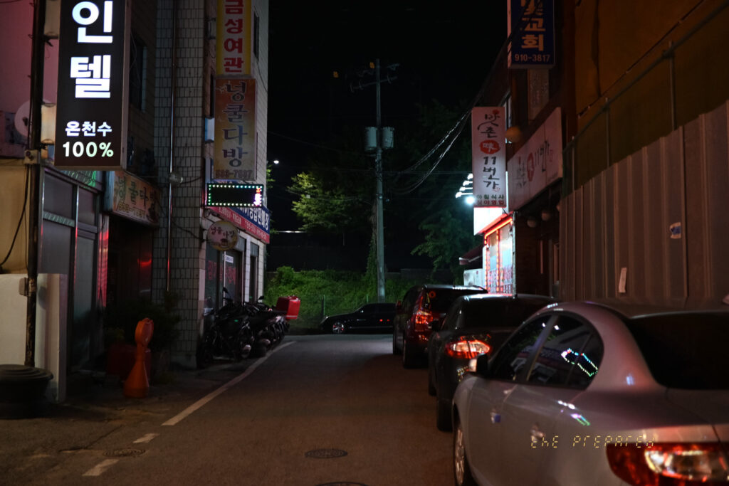 아산시의 어두운 밤거리를 담은 사진입니다. '인텔 온천수 100%'라고 적힌 간판이 있는 건물, 빨간색 여관 간판, '덩굴 다방'이라고 적힌 간판, 그리고 교회 간판이 나란히 보입니다. 좁은 길가에는 몇 대의 오토바이와 자동차가 주차되어 있고, 가로등 불빛이 희미하게 거리를 비추고 있습니다.