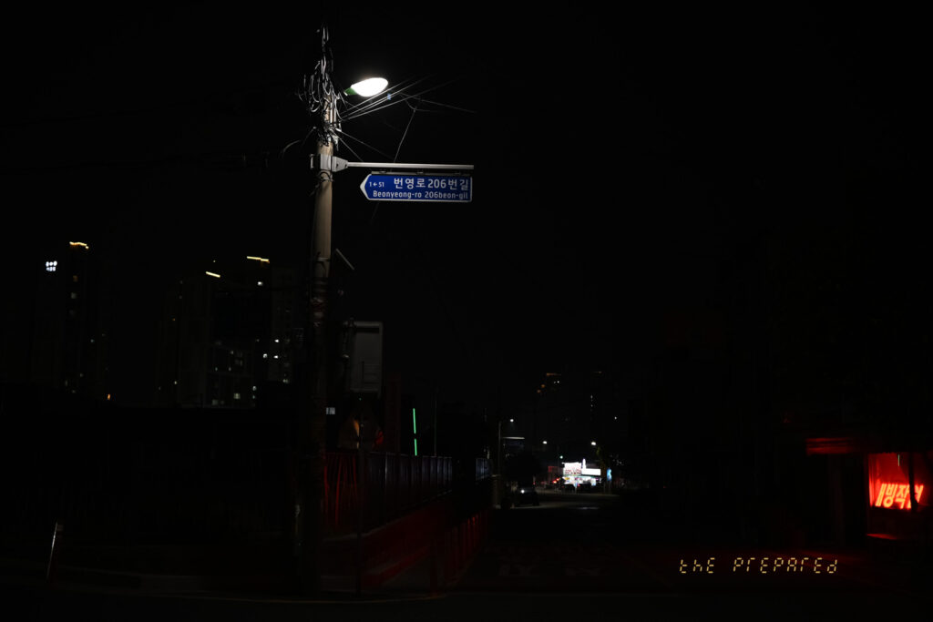 검은 배경에 파란색 거리 표지판과 희미한 가로등 불빛이 보이는 밤거리 사진.