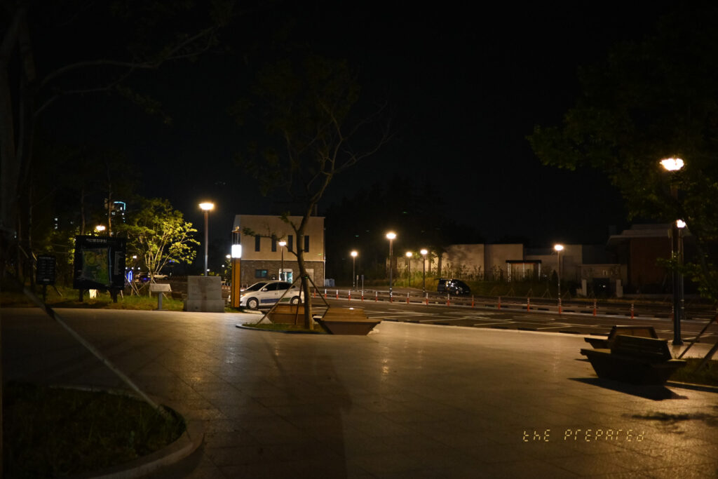 밤의 주차장 이미지는 불이 켜진 도로와 건물, 벤치가 있는 어두운 장면을 보여줍니다.