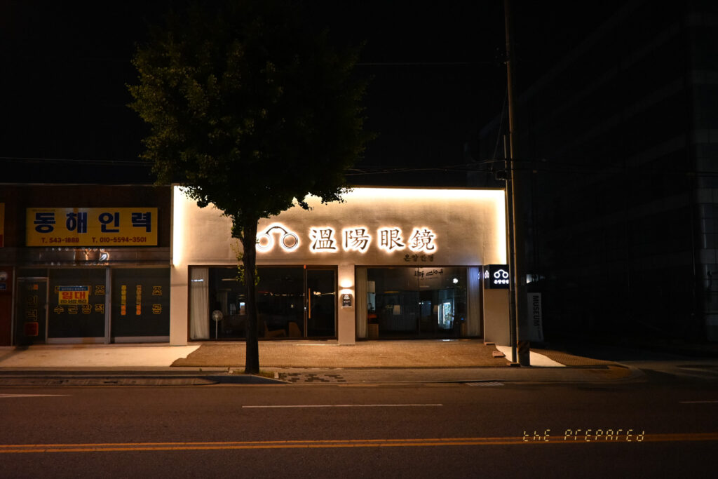 어두운 밤거리에 텅 빈 상점 건물 앞을 담은 이미지. '온양안경' 간판에 은은한 빛이 비추고 있으며, 건물 옆에 가로등과 나무 한 그루가 서 있습니다.
