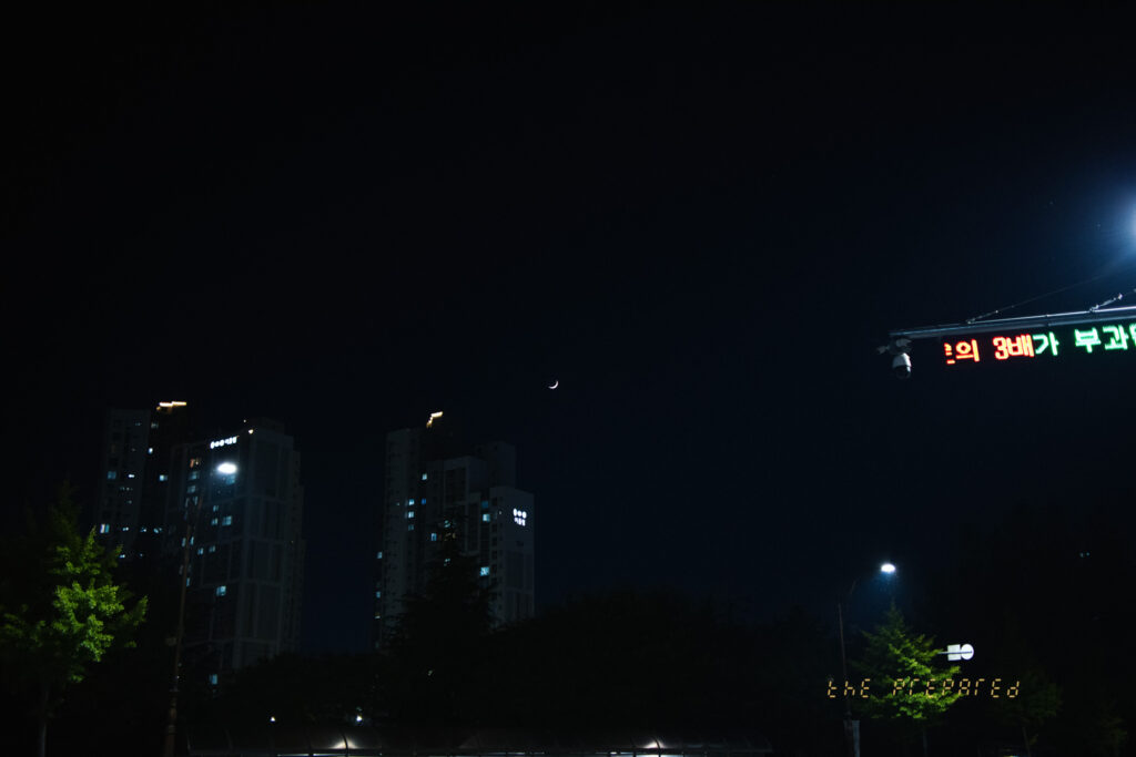 어두운 배경에 도시의 건물들과 가로등, 그리고 초승달이 보이는 밤하늘 사진입니다. 오른쪽에는 LED 간판의 일부가 보이며, 텍스트의 마지막 두 글자가 부분적으로 드러나 있습니다.