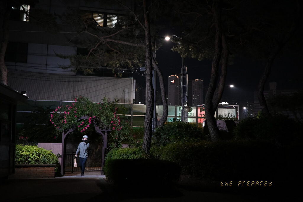 저녁에 한 남자가 도시의 길을 걷는 모습을 보여줍니다. 어두운 배경과 가로등 불빛 속에서 남자는 눈에 띄게 대비됩니다. 일부 식물과 꽃이 배경에 나타나 이미지에 약간의 색을 더합니다.