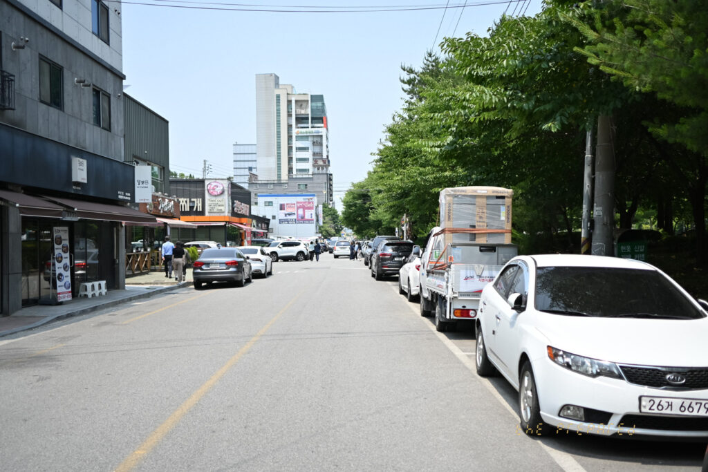 아산시의 한적한 도로 풍경 사진. 2013 기아 K3 흰색 차량이 도로 옆에 주차되어 있고, 맞은편에는 다양한 상점들이 늘어서 있습니다.