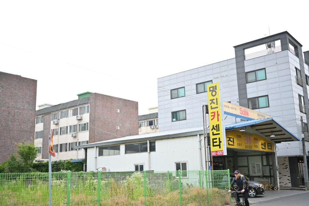 주거 지역의 자동차 정비소 사진, 도로 맞은편에는 다양한 건물이 보이고, 한 남자가 자전거를 타고 건물 앞 도로를 지나가고 있습니다