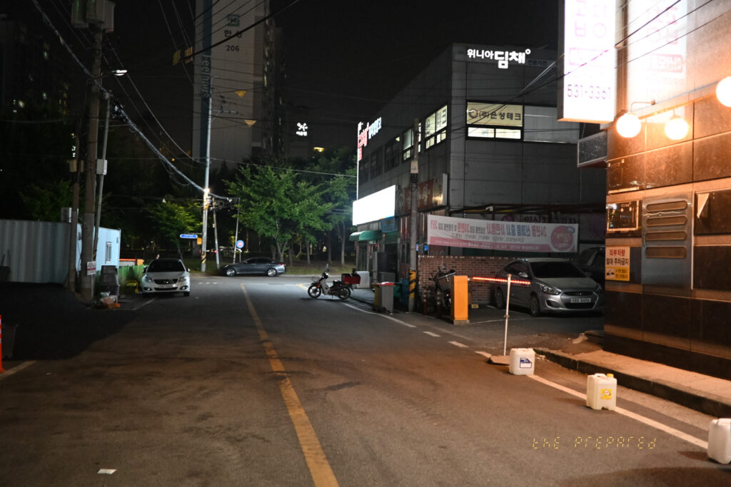 아산시의 어두운 밤거리를 담은 사진입니다. '위니아 딤채' 간판이 걸린 건물과 주차된 차량 몇 대, 그리고 오토바이 한 대가 보입니다. 좁은 골목길에 가로등 불빛이 희미하게 비추고 있습니다.