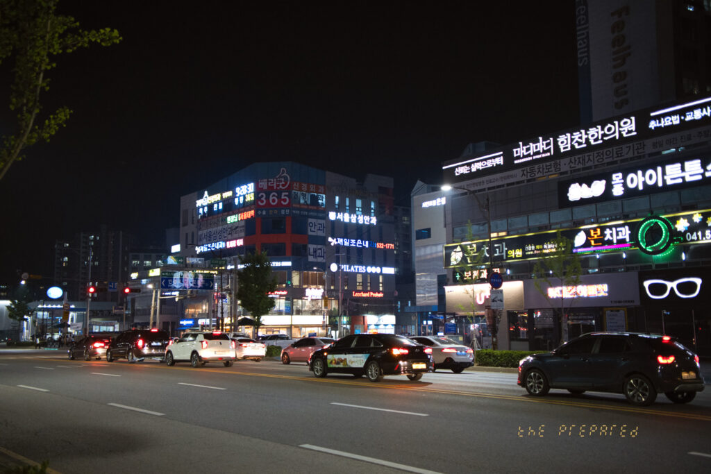 야간 조명이 켜진 아산시 번화가 도로 풍경 사진. 여러 대의 자동차가 도로를 따라 늘어서 있고, 다양한 간판과 상점들이 눈에 띕니다. 오른쪽에는 렌즈 모양 간판과 안경 그림이 크게 보입니다.