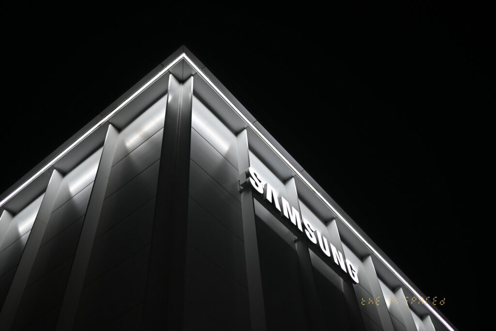 검은색과 흰색의 극적인 사진. 건물의 아래쪽에서 촬영하여 아래쪽에서 위쪽을 향하게 보여줍니다. 건물은 검은색 배경에 흰색 선으로 강조된 건축적 모서리와 각도를 나타냅니다. 이미지의 주요 초점은 어두운 환경과 뚜렷한 대조를 이루는 건물 모서리에 부착된 조명으로 강조된 "SAMSUNG"이라는 단어입니다. 미적 측면은 흑백 색상과 건물 디자인으로 강조되는 건축 자재를 강조합니다.