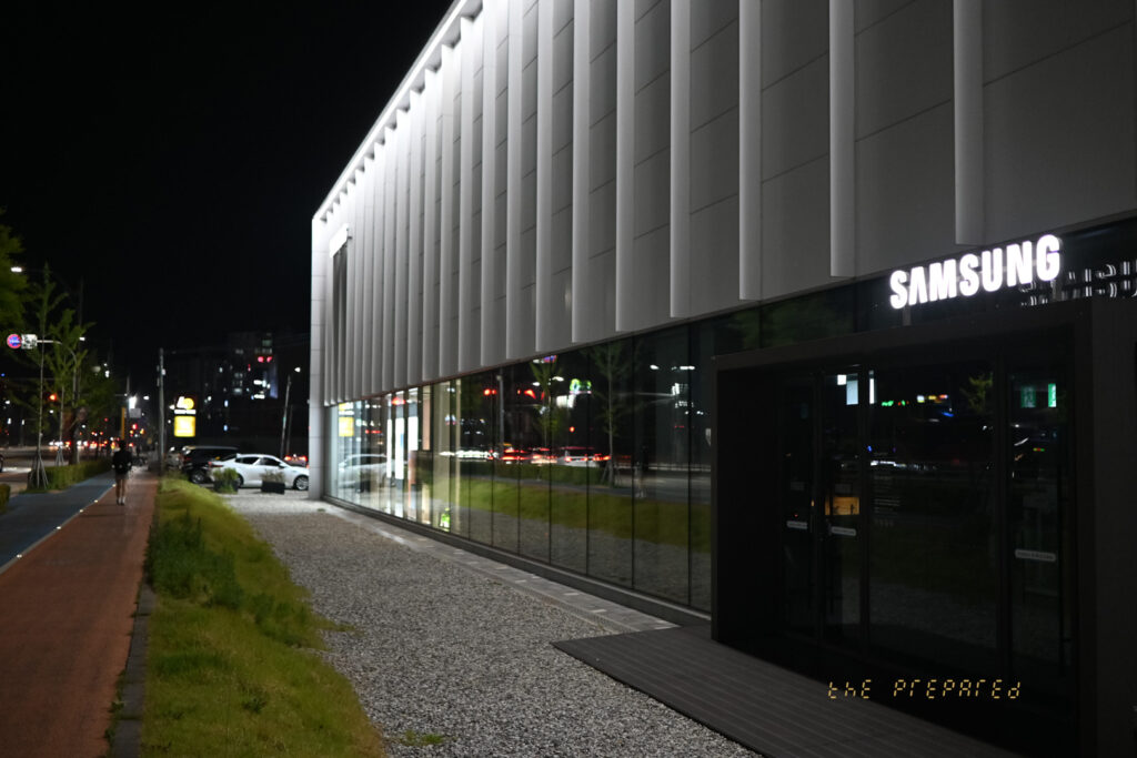 저녁에 건물 외부에서 찍은 삼성 매장 사진. 건물의 외벽은 회색과 흰색 줄무늬로 덮여 있으며 큰 삼성 로고가 건물 앞면에 있습니다. 매장 앞에는 포장된 보도가 있습니다.