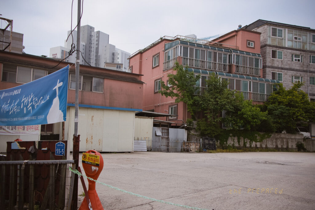 한국 아산시의 주거 지역을 담고 있습니다. 분홍색 건물이 주요 피사체이며, 울창한 나뭇가지가 건물을 둘러싸고 있습니다. 전선과 전봇대가 프레임을 가로지르며, 도로 옆에는 주차 금지 표지판이 세워져 있습니다. 55-5라고 적힌 파란색 표지판이 있는 십자가 배너도 볼 수 있습니다. 회색의 우울한 날씨는 도시 외곽의 고요하고 인적이 드문 장면을 연출합니다.