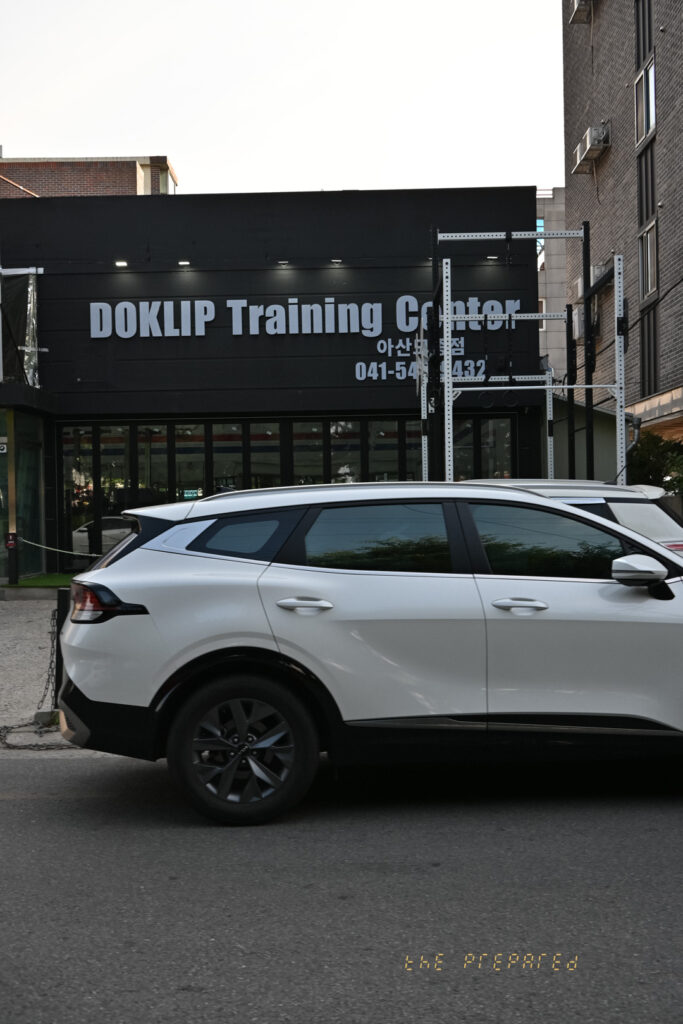 아산에 있는 "DOKLIP Training Center"라는 검은색 간판의 피트니스 센터 건물 사진. 건물 앞에 2023 기아 스포티지 LX 흰색 차량이 주차되어 있습니다.