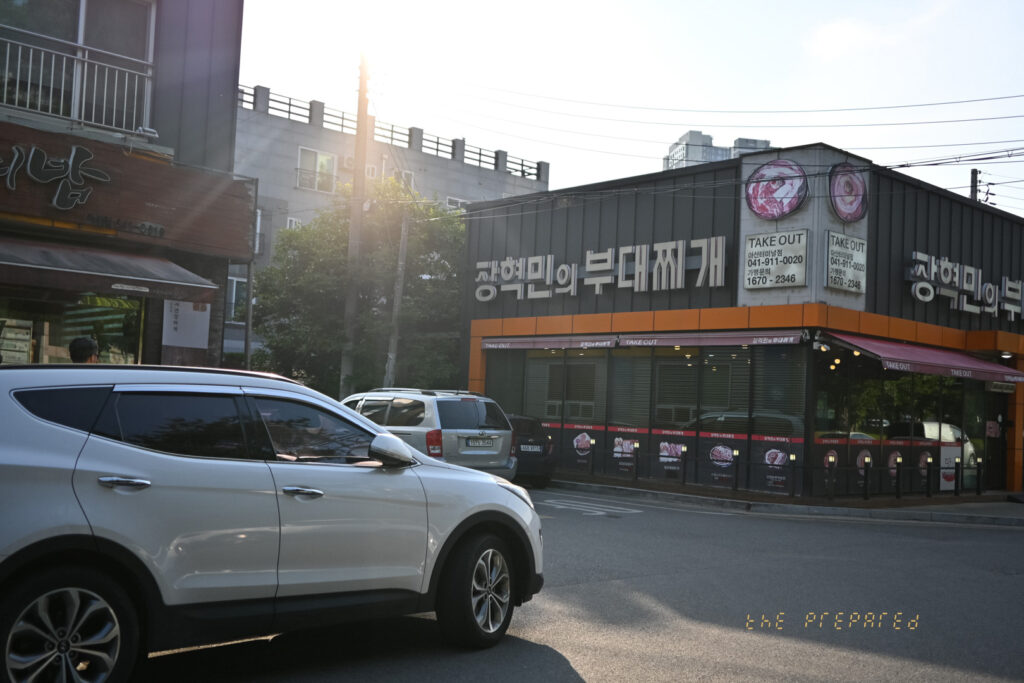 충청남도 아산의 "장혁민의 부대찌개"라는 음식점의 바깥쪽 사진입니다. 이 음식점은 검은색과 주황색이고, 도로에서 몇 대의 차량을 볼 수 있습니다. 오른쪽에는 다른 건물이 보입니다.