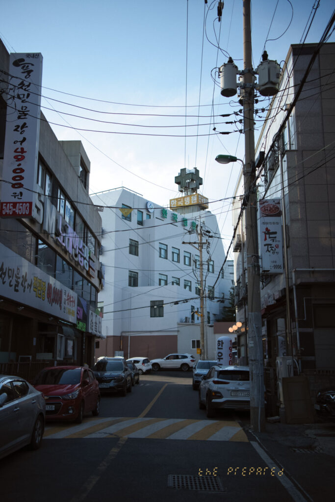 아산시 도심의 좁은 골목길을 담은 사진. 전봇대와 복잡하게 얽힌 전선이 보이고, 양옆에는 상가 건물이 늘어서 있습니다. 골목길 안쪽에는 하얀색 건물이 보이며, 골목길에는 차량 몇 대가 주차되어 있습니다.