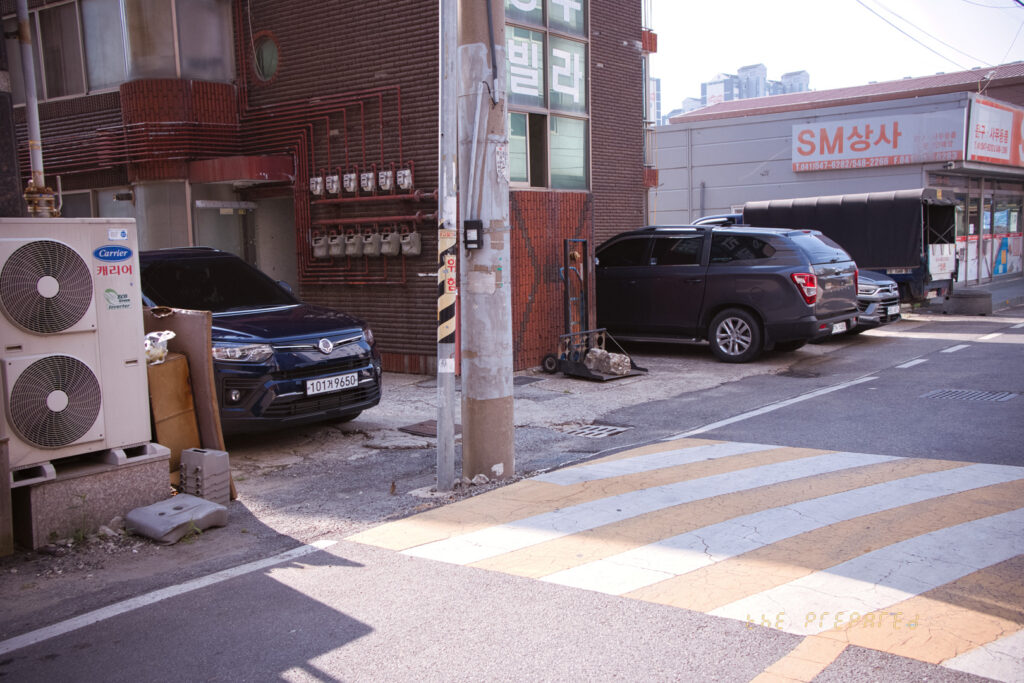 한국 충청남도 아산에 있는 동네의 이미지입니다. 다양한 회사의 자동차들이 있습니다. 여기에는 검은색 현대 투싼과 짙은 회색 쌍용 렉스턴 스포츠가 포함됩니다. 에어컨 장치가 한쪽에 있고, 차량은 건물을 향하고 있으며, 거리 반대편에 있는 또 다른 건물은 SM 상사입니다.