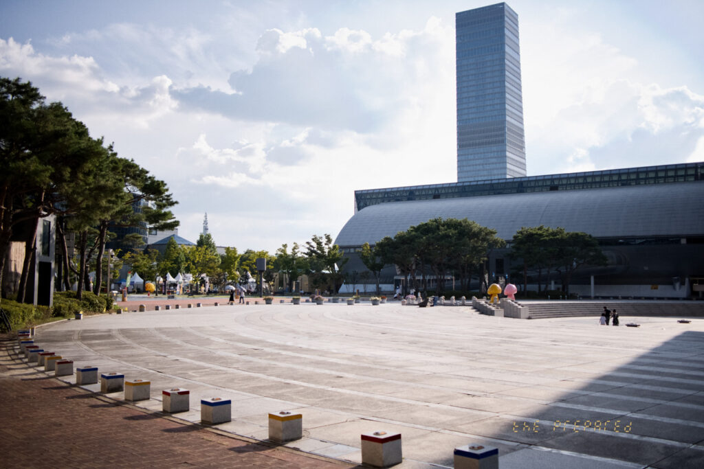 탁 트인 광경의 사진으로, 넓은 광장과 몇 개의 나무, 그리고 키가 큰 건물을 볼 수 있습니다. 구름과 햇빛이 있는 밝은 날로, 사람들이 광장 주변을 거닐고 있습니다.