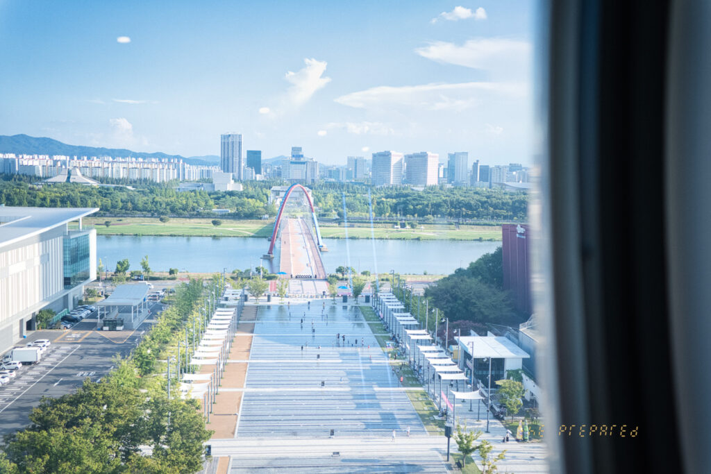 창문을 통해 바라본 엑스포 음악 분수 공원의 전경 사진입니다. 긴 산책로와 다채로운 아치형 다리, 아름다운 도시 풍경을 볼 수 있습니다.