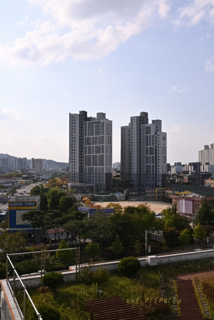 푸른 하늘 아래 2개의 고층 아파트 단지가 보이는 아산시 풍경 사진. 1층 건물과 나무들이 도시 풍경을 더욱 돋보이게 합니다.