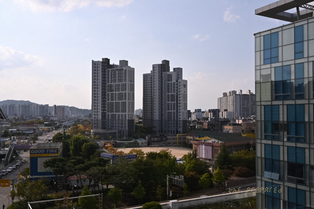 푸른 하늘 아래 2개의 고층 아파트 단지가 보이는 아산시 풍경 사진. 1층 건물과 나무들이 도시 풍경을 더욱 돋보이게 합니다.